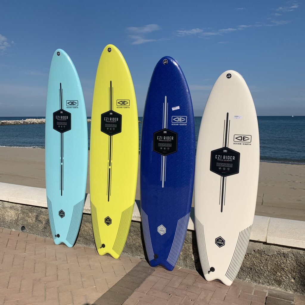 Diversas tablas de surf para principiantes de Dreisog expuestas ante la playa: colores azul clarito, azul oscuro, amarillo y blanco.