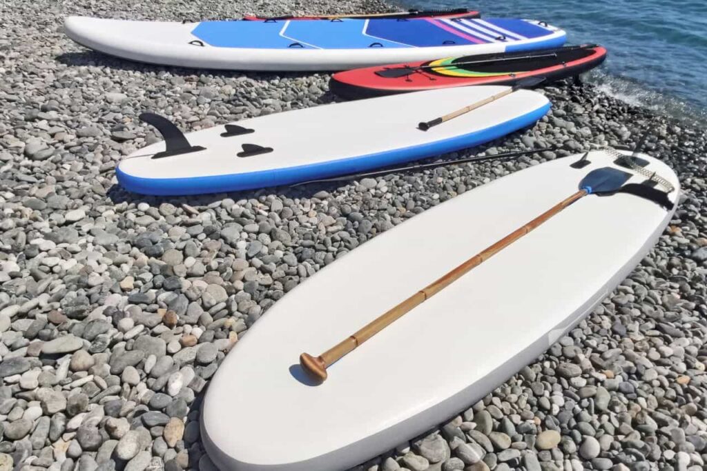 Distintas tablas de paddle surf y remos sobre playa de rocas.