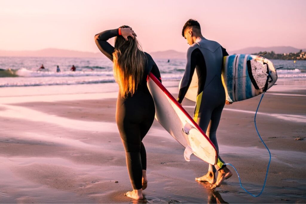 Mujer y hombre surferos acercándose a la orilla del mar, ambos dos portando tablas de surf con las medidas de las tablas de surf acordes a su peso y altura.